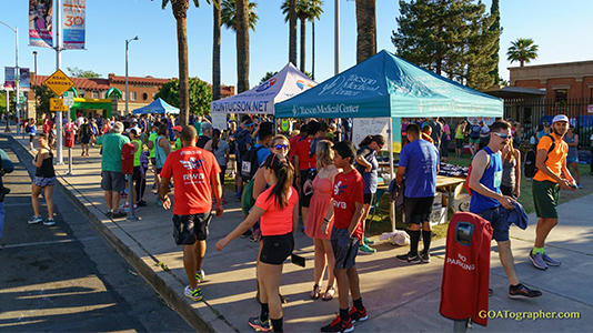 Outdoor event with people in athletic attire walking and standing near tents, including ones labeled "Tucson Medical Center" and "Run Tucson," on a sunny day.