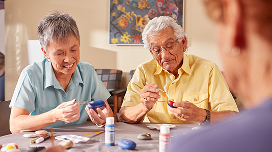 Elderly individuals painting rocks at a table covered with painting supplies.