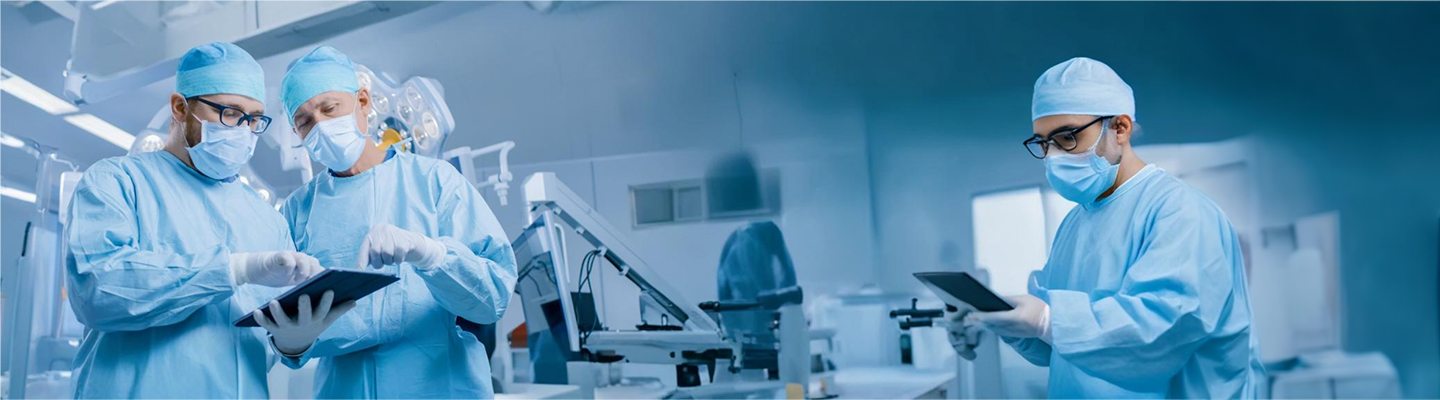 Three medical professionals in blue surgical attire examining tablets in a hospital setting.