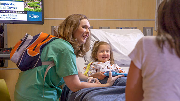 A smiling young girl in a hospital bed with a toy, a woman in green scrubs with a backpack leaning towards her, and another person in a white top.