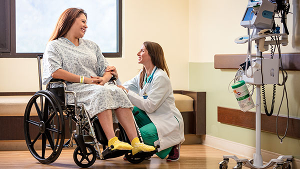 A patient in a wheelchair and a healthcare professional smiling and talking in a hospital room.