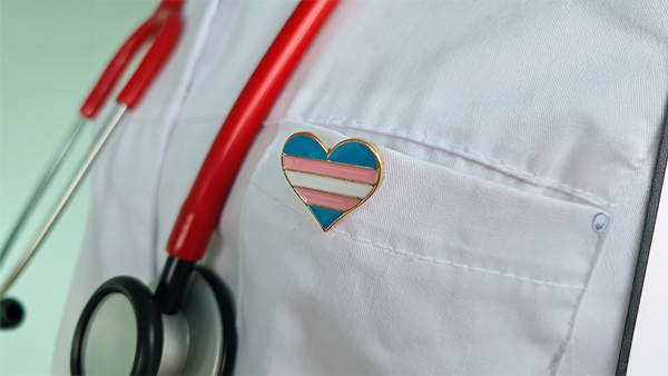 A white lab coat with a heart-shaped transgender pin and a red stethoscope.