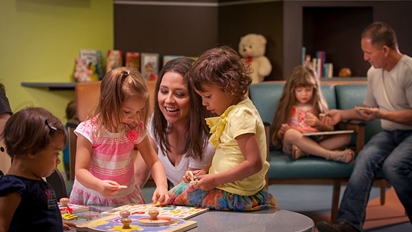 Children playing with toys at a table in a colorful playroom with adults supervising in the background.