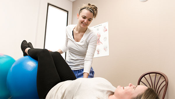 A standing woman assists another woman lying on a blue exercise ball in a clinical setting.