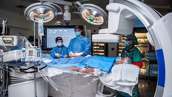 Medical professionals performing surgery with overhead lights, monitors, and medical equipment around the patient.