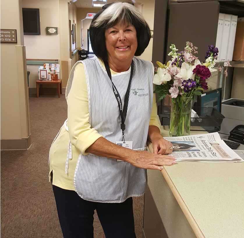 A hosice volunteer in a striped apron stands next to the front desk and smiles at the camera.