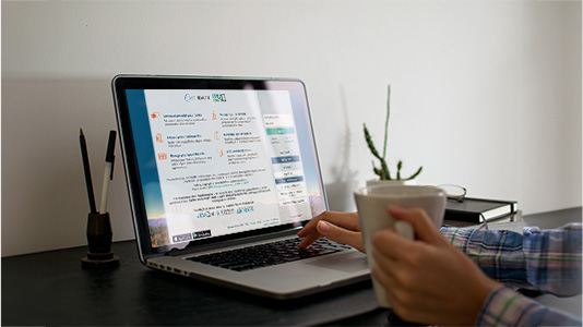 Person with a plaid shirt holding a mug while logging in TMC MyChart in a laptop on a desk with a small potted plant and a notebook.