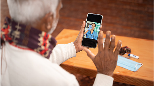 Elderly person on a video call with a healthcare professional, holding a smartphone. A table in the background has a blue surgical mask, a blister pack of pills, and a white inhaler.