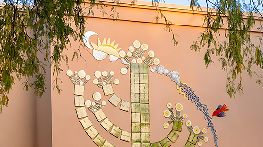 A wall adorned with a colorful mosaic artwork featuring abstract, branching shapes resembling a tree or plant with round, flower-like elements. The design includes a mix of gold, green, and white tiles, with a sun and moon depicted at the top left, and a small red bird flying toward the right. Green tree branches hang down into the frame from the top.