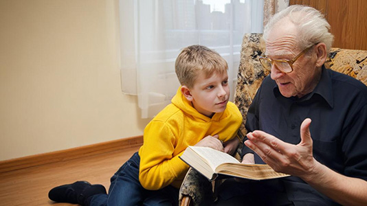 An older man sits in a comfortable chair with a tween boy lounged on the floor next to him. The man is reading out of a thick, old book to the boy.