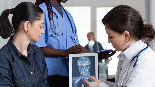 A medical professional, wearing a white coat and stethoscope, shows a patient a digital image of a neck and brain scan on a tablet. The patient, dressed in a dark shirt, looks attentively at the screen.