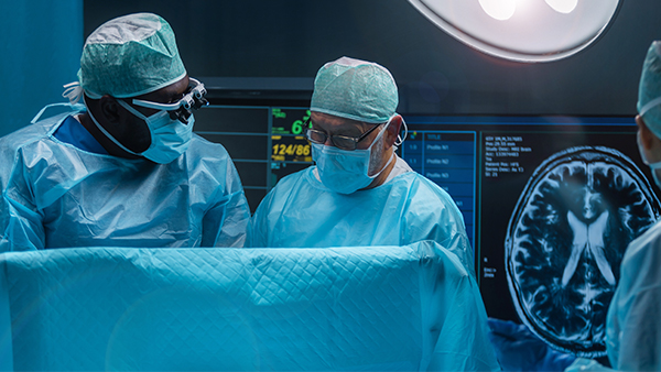 Two surgeons in an operating room with a brain scan displayed on a monitor.