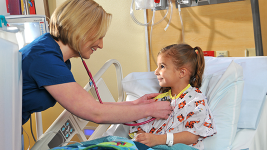 a medical professional use a stethoscope on a smiling child in a hospital bed