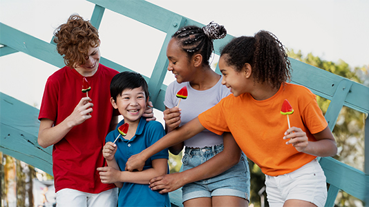 Four preteen kids holding watermelon ice cream on sticks, standing together outdoors, and smiling.