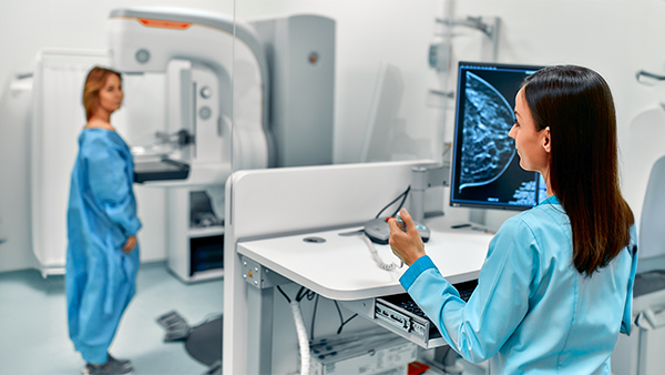 Medical professional reviewing a mammogram on a computer as a patient stands near a mammography machine.