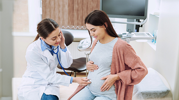 A healthcare professional listens to a pregnant woman's belly using a stethoscope in an examination room.