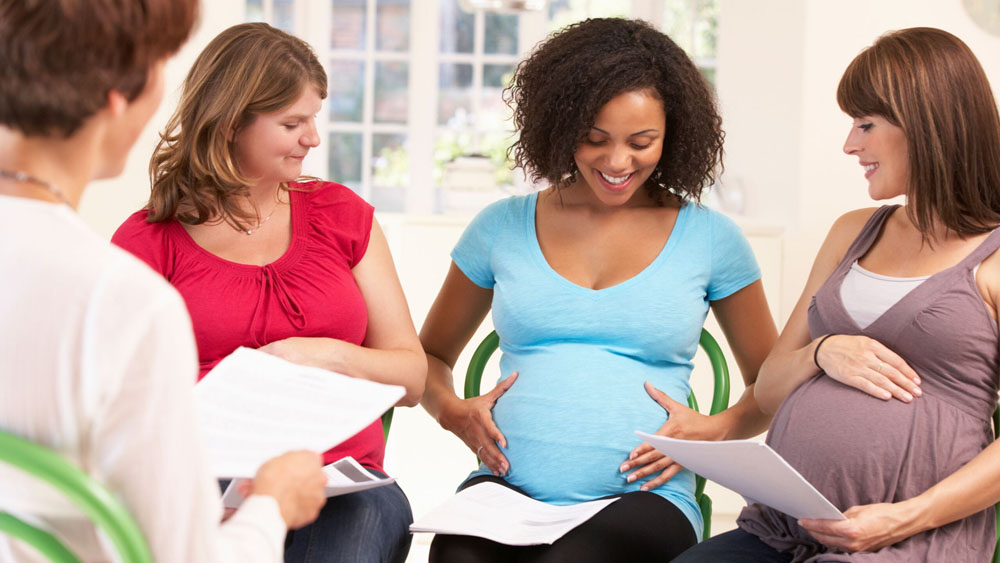 Group of three pregnant women sit together with hands on their own stomach and gaze at the belly of central expectant mother